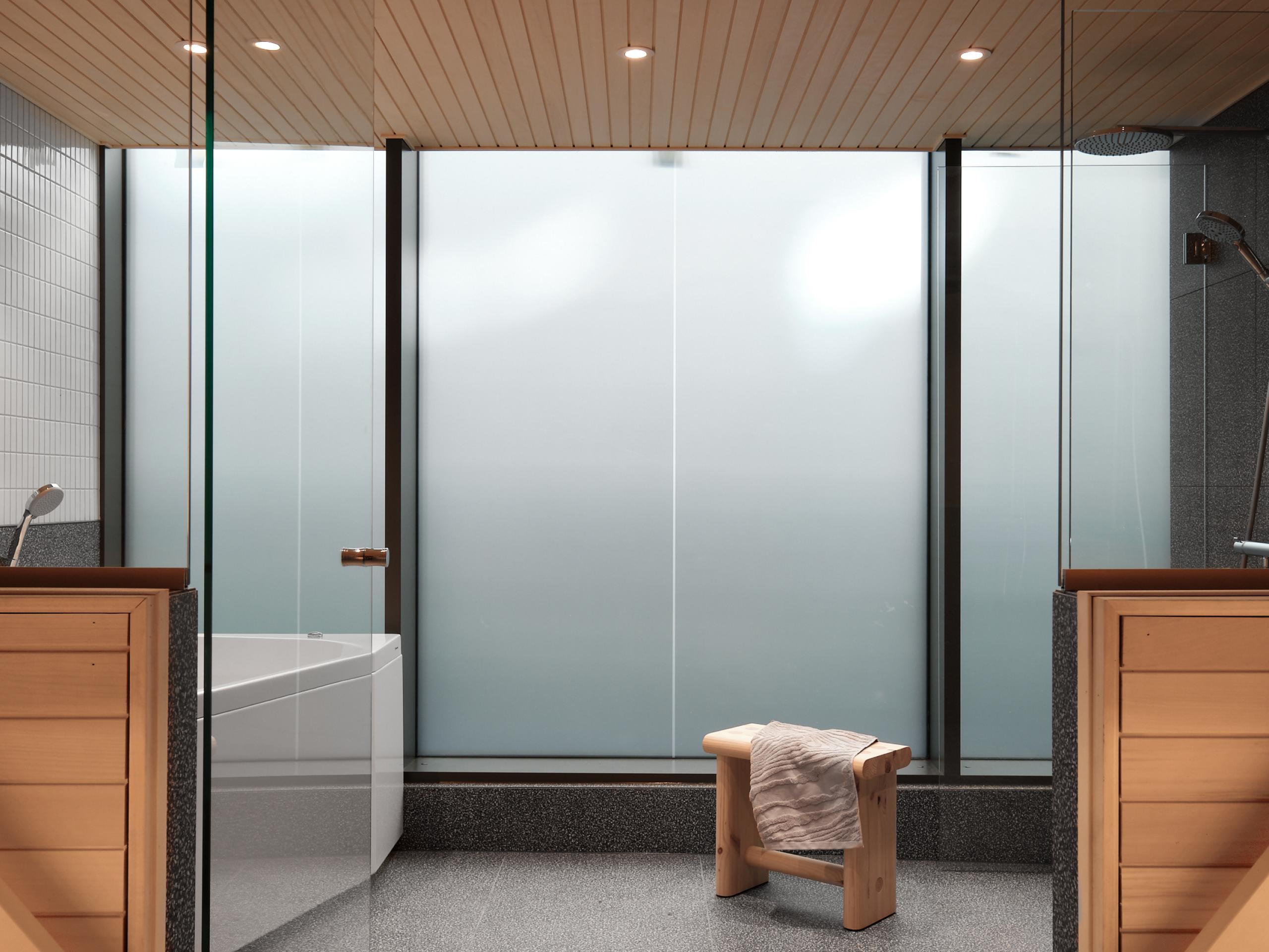 Suihkutila, jossa on himmeät lasi-ikkunat, vaaleat puuyksityiskohdat ja puinen jakkara, mikä luo rauhallisen ja yksityisen ilmapiirin.