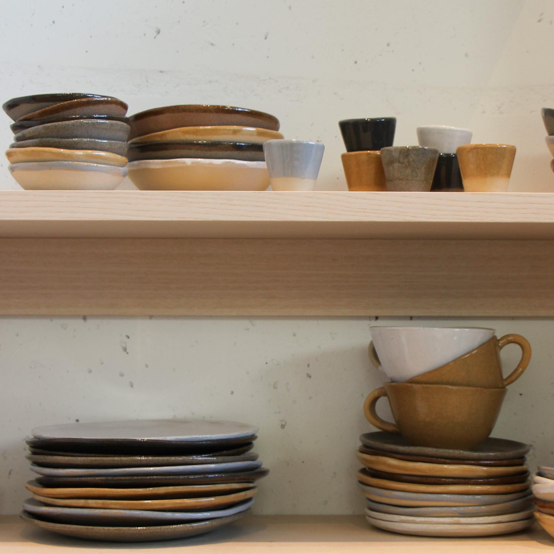 Kuvassa on Raaka Rå -keramiikkastudion käsintehty astiasto, joka sisältää valikoiman lautasia, kulhoja, kuppeja ja pieniä astioita maanläheisissä sävyissä, kuten ruskea, harmaa ja beige. Keramiikka on minimalistista ja rustiikkista, matta- ja kiiltävillä pinnoilla, aseteltuna siististi puuhyllyille.