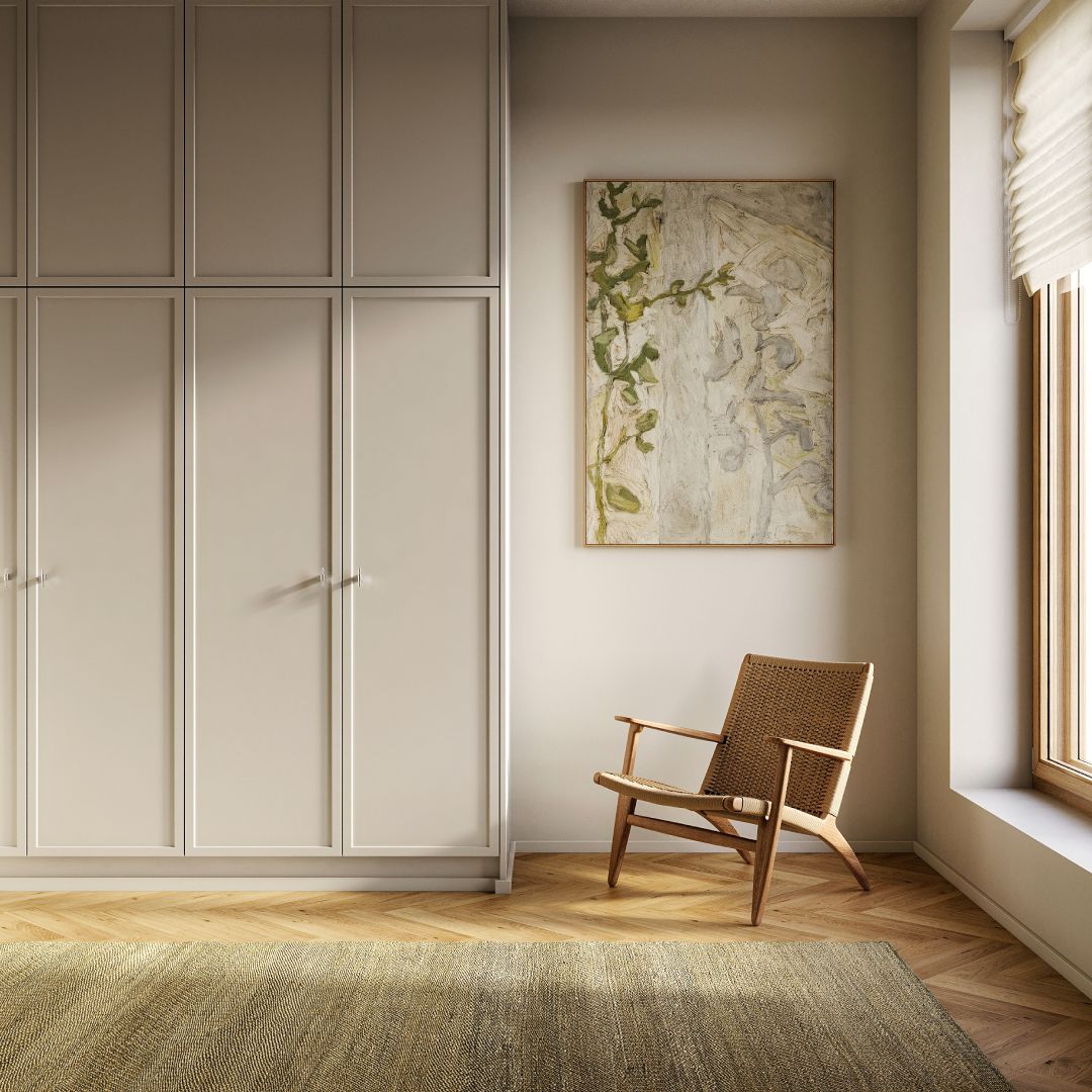 Vaalea, minimalistinen huone, jossa on suuri seinän levyinen kaapisto, parkettilattia kalanruotokuviolla, seinällä abstrakti maalaus sekä ikkunan vieressä puinen nojatuoli punotulla istuimella. Luonnonvalo tulee sisään oikealla olevasta ikkunasta.