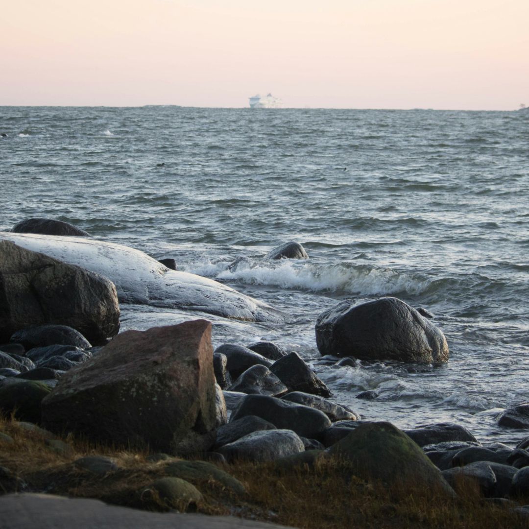 Kivikkoinen Itämeren ranta hämärässä. Suuret graniittikivet laskeutuvat aallokoiseen mereen, taustalla vaaleanpunainen iltataivaanranta. Horisontissa näkyy alus.